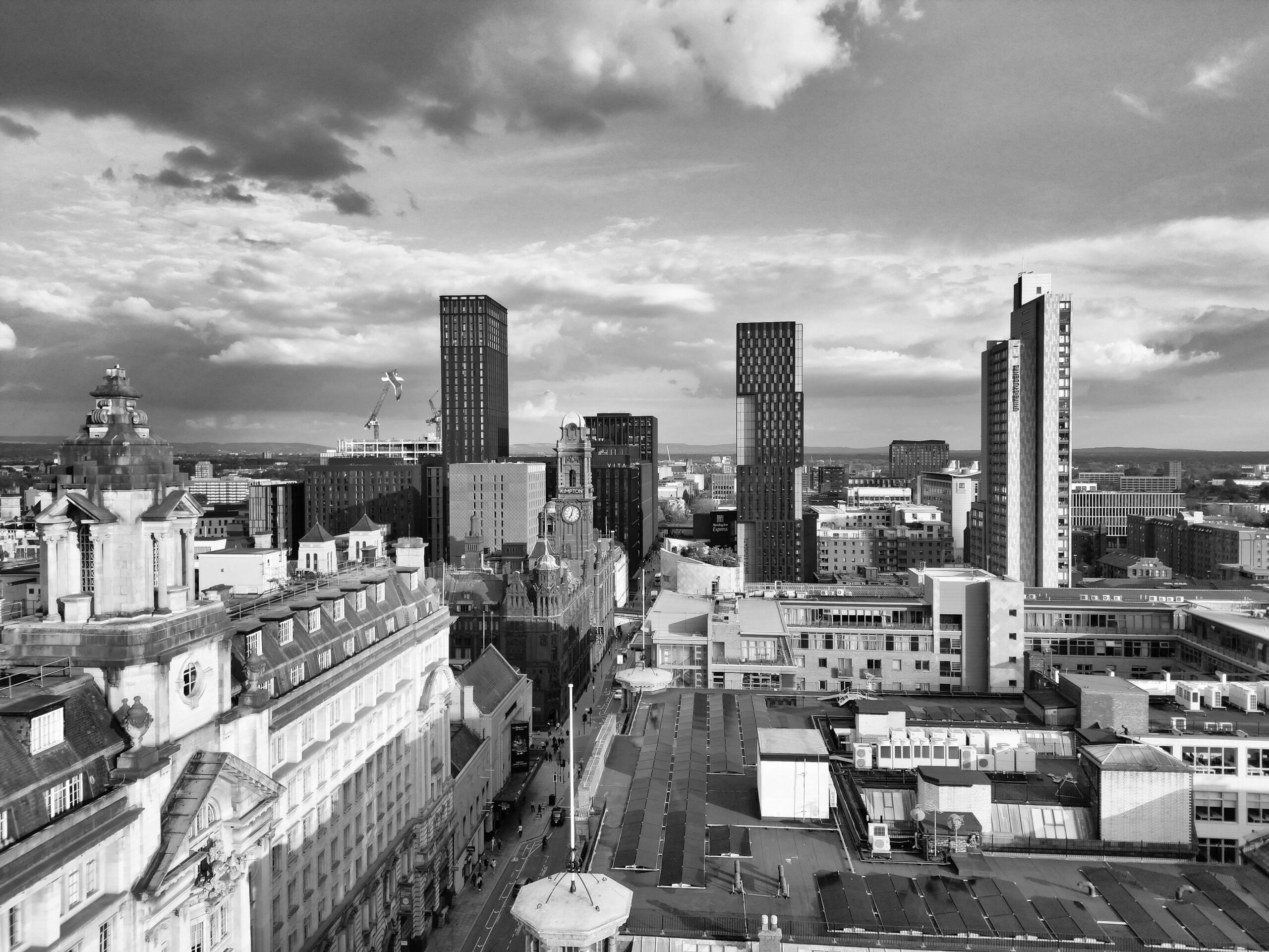 A dramatic aerial view of Manchester's modern skyline under a cloudy sky in black and white.